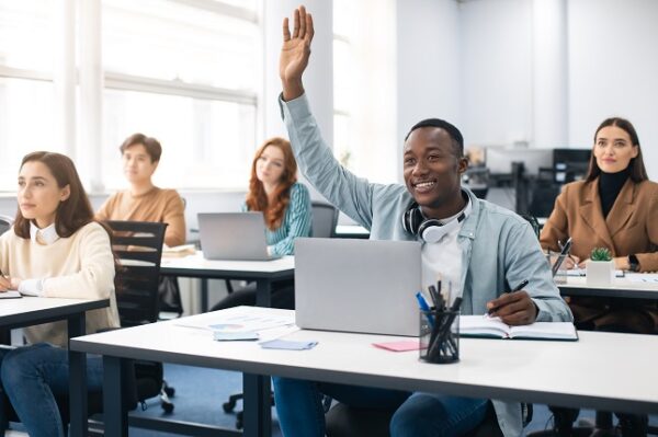 Education Concept. Smiling African American Male Student Raises Hand Up and Asks Lecturer a Question, Using Laptop Listening to Lecture at the University. Multi Ethnic Group of Modern Bright Students